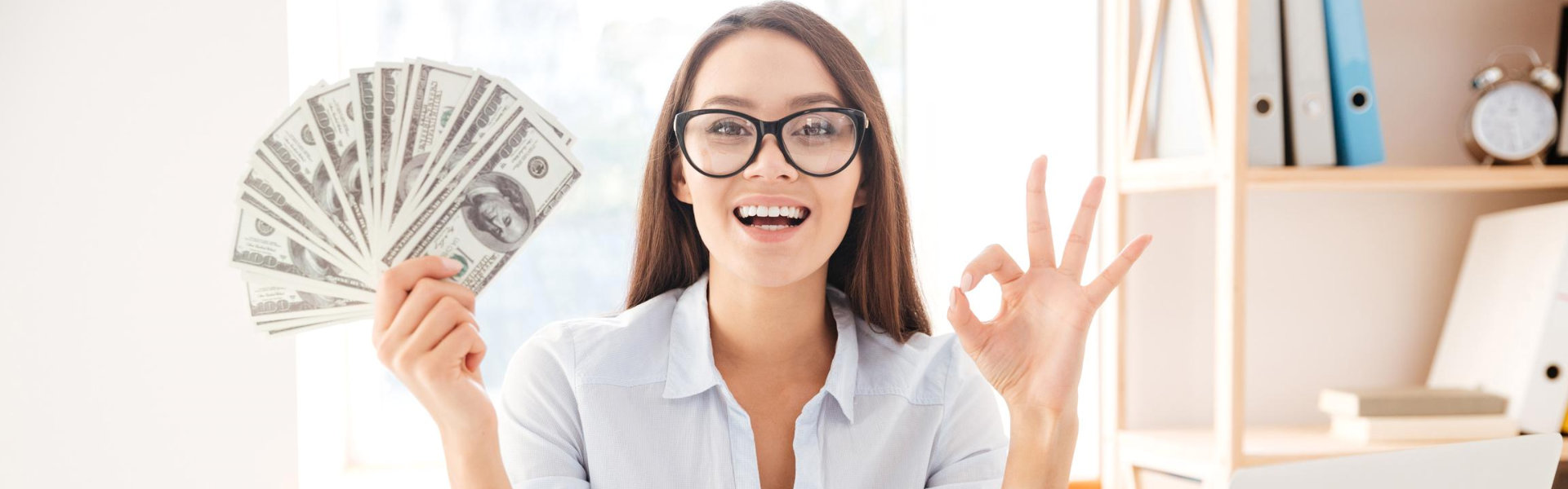 Image of businesswoman dressed in white shirt sitting in her office and holding money in hand while making Okay gesture