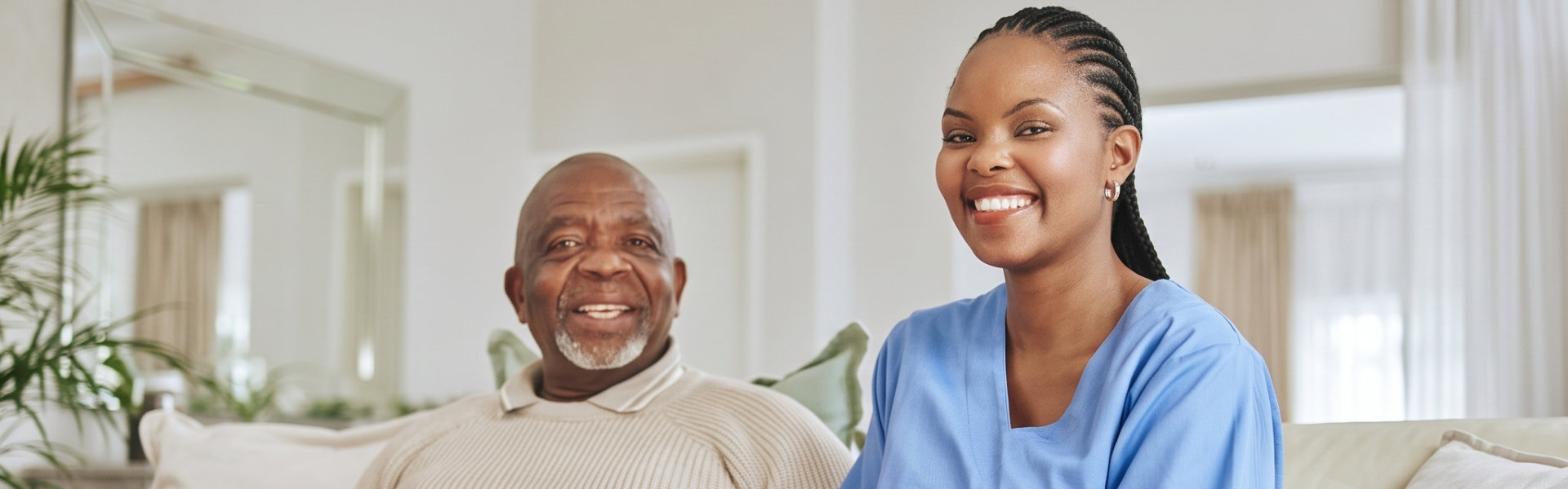 A smiling nurse holding hands with an elderly man on a couch in a living room.