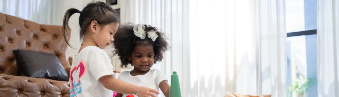 Two young children, one with dark hair in a ponytail and the other with dark, curly hair adorned with a large white bow, are playing together indoors, interacting with a toy.