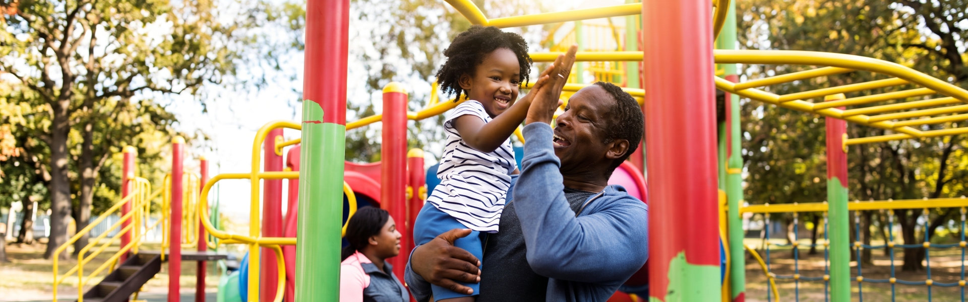a smiling man in a blue hoodie holds up a young child with curly hair wearing a striped shirt